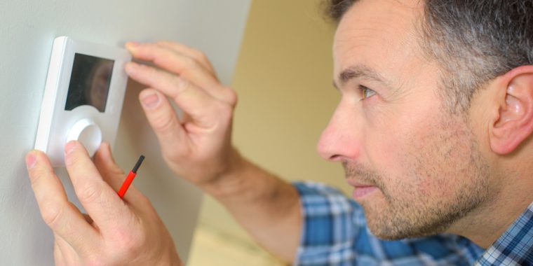 A man with a screwdriver inspects a wall-mounted thermostat